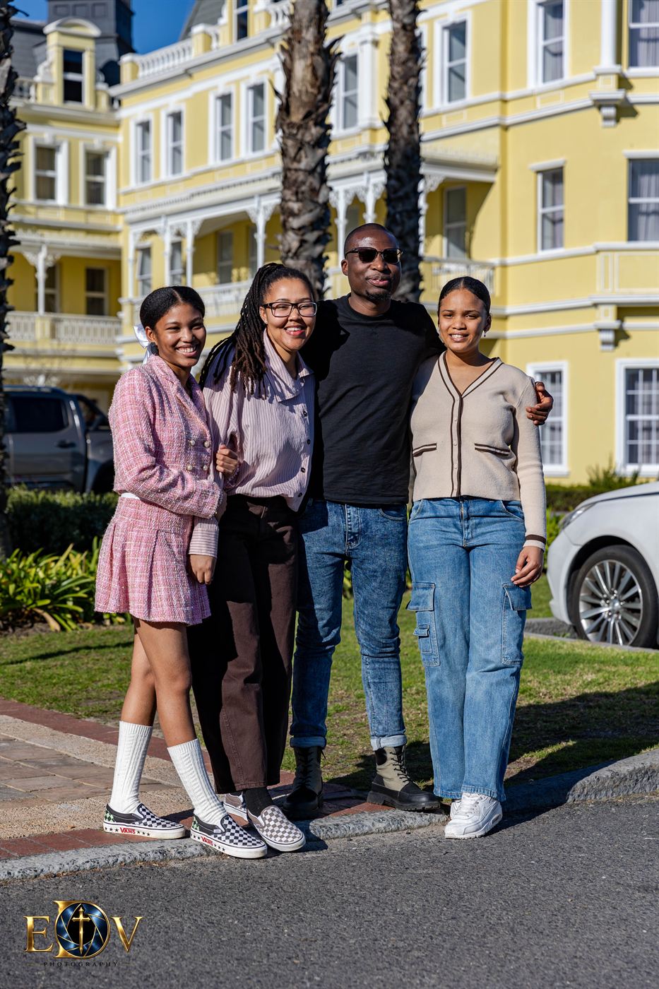 Group of four standing arm in arm outside a yellow Victorian building lined with palm trees