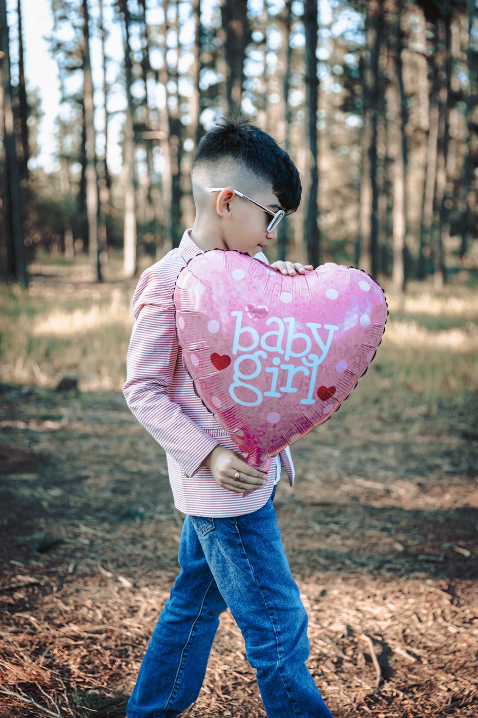 Young boy carrying a pink heart balloon reading baby girl through a pine forest