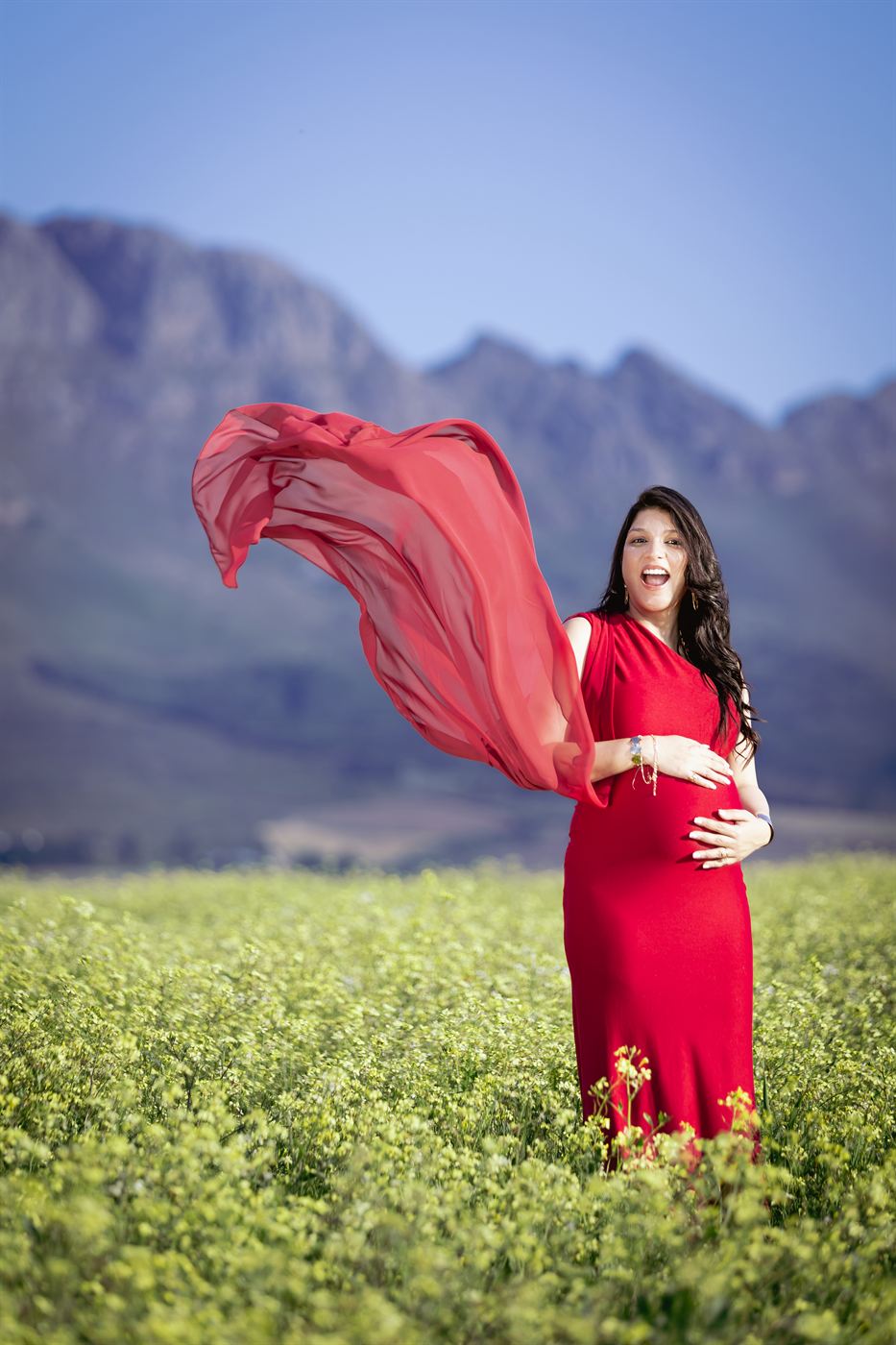 Expectant mother in a red gown, her scarf caught by the wind in a flowering field