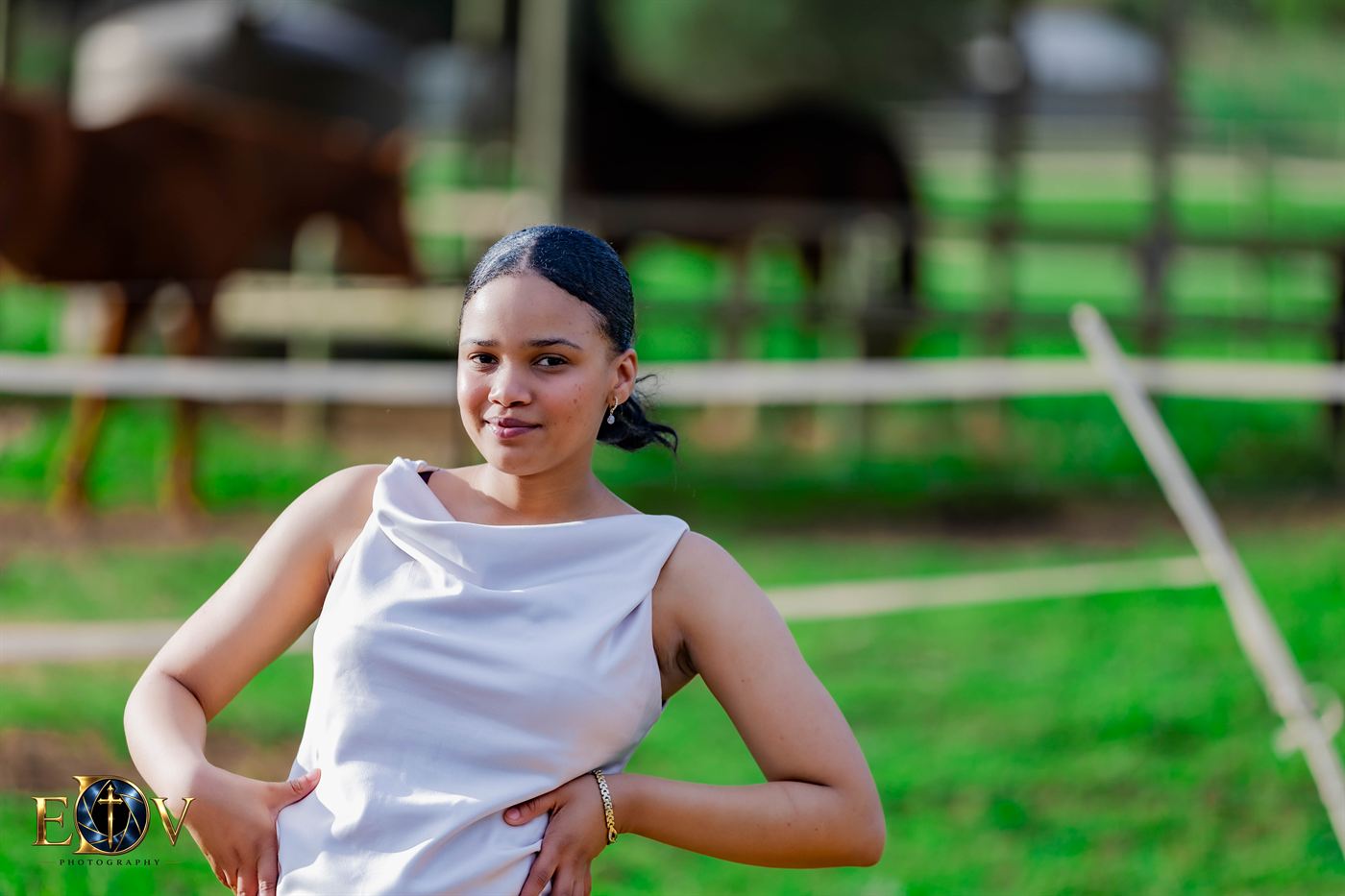 Young woman in a satin top standing beside a paddock, horses grazing behind her