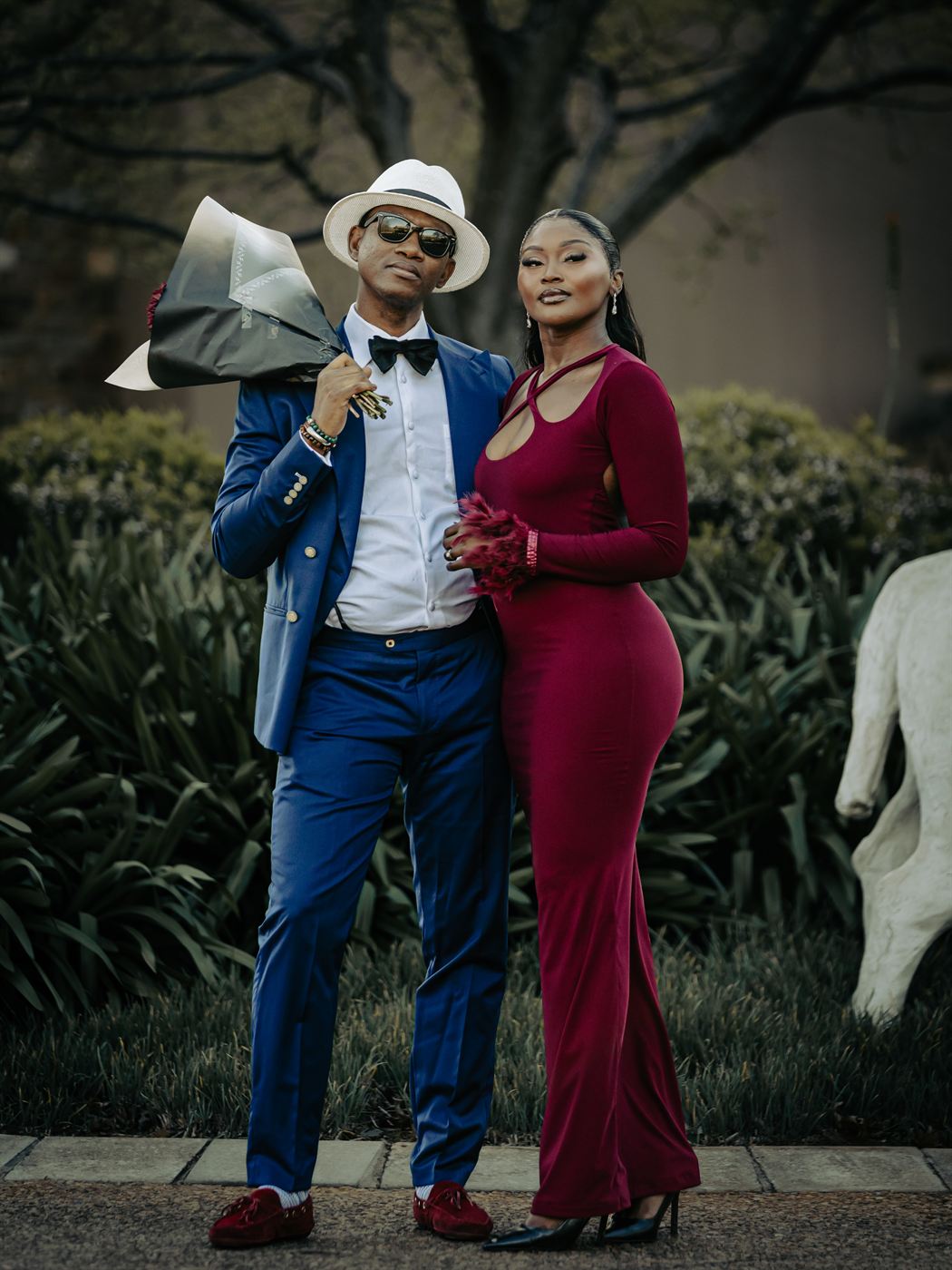 Couple posing in a garden, he with a bouquet over his shoulder, she in a deep red gown
