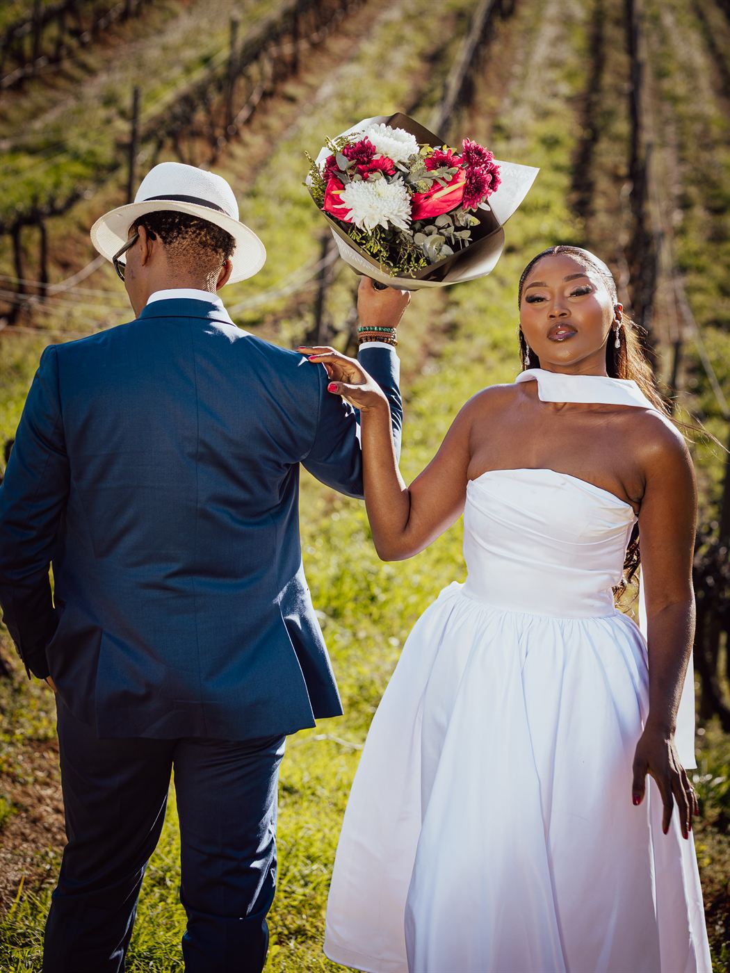 Groom holding a bouquet aloft in a vineyard as the bride reaches for it